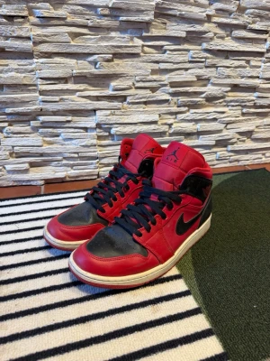 Nike Air Jordan 1 Mid röd/svart - Säljer ett par Nike Air Jordan 1 Mid sneakers i klassisk röd och svart färg med vita sulor. Skorna har svart swoosh, svarta skosnören och är tillverkade i läder. Perfekta för dig som gillar streetwear och ikoniska sneakers.