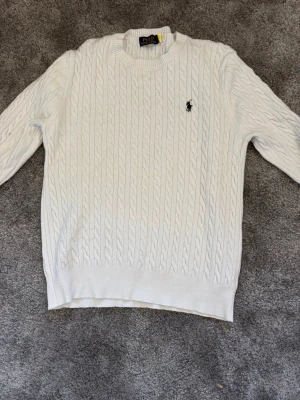 Vit kabelstickad tröja Polo Ralph Lauren - Vit kabelstickad tröja från Polo Ralph Lauren med rund halsringning och broderad logga på bröstet. Tröjan har långa ärmar och ribbade muddar vid ärmslut och nederkant. Perfekt för en klassisk och stilren look.