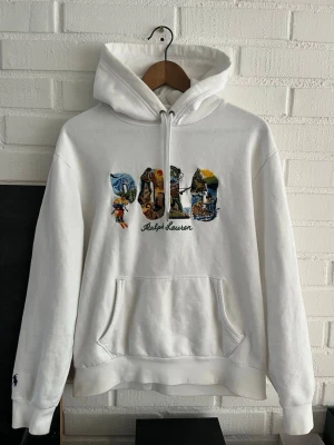 Vit hoodie från Ralph Lauren - Säljer en vit hoodie från Ralph Lauren med stor broderad POLO-text på bröstet fylld med färgglada outdoor-motiv. Hoodien har huva med dragsko, magficka och ribbade muddar. Perfekt för dig som gillar streetwear och vill sticka ut med snygga detaljer.