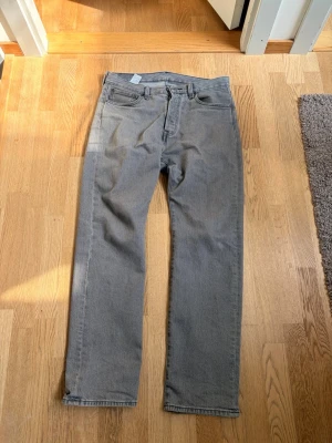 Grå raka jeans från Levi's (32:32) - Säljer ett par grå jeans från Levi's med klassisk femficksdesign och raka ben. Jeansen har en diskret tvätt och Levi's ikoniska läderpatch bak i midjan. Perfekta för en avslappnad och stilren look.