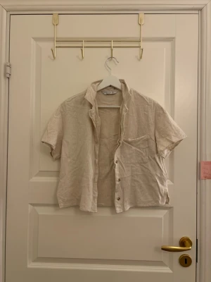 Beige linneskjorta från Stradivarius - Kortärmad beige skjorta från Stradivarius i luftig linnemix. Skjortan har klassisk krage, knappstängning framtill och en bröstficka. Perfekt för en avslappnad och stilren look.