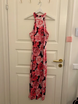 Lång blommig klänning med rosor - Snygg långklänning med stora röda och rosa rosor på svart botten. Klänningen har hög hals och är ärmlös, vilket ger en modern och fräsch look. Perfekt för dig som vill sticka ut med ett färgstarkt mönster. Materialet känns mjukt och följsamt.