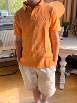 Orange pikétröja från Polo Ralph Lauren - Snygg orange pikétröja från Polo Ralph Lauren med klassisk krage och korta ärmar. Tröjan har en liten blå broderad logga på bröstet och är tillverkad i mjuk bomull. Perfekt för en avslappnad och stilren look.