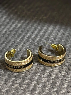 Guldiga örhängen från Christian Dior - Snygga guldiga örhängen från Christian Dior med gnistrande stenar och svart detalj med märkets namn. Halvbågsformade och eleganta, perfekta för att lyfta din stil. Klassisk design med exklusiv känsla.