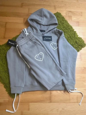 Grått loungewear-set från Carroza - Mysigt grått loungewear-set från Carroza med hoodie och matchande byxor. Båda plaggen har ett vitt hjärta med ansikte broderat på bröstet och benet. Hoodien har känguruficka och dragsko i huvan, byxorna har resår och dragsko i midjan.