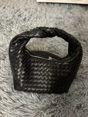 Svart flätad handväska i skinn - Trendig svart handväska i flätat skinn med silvrig dragkedja och rundad form. Väskan har ett mjukt handtag och rymligt fack, perfekt för att bära dina viktigaste saker. Snygg och stilren accessoar som lyfter din outfit.