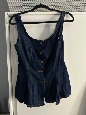 Mörkblå denim topp med knappar - Snygg mörkblå denim topp med breda axelband och dekorativa metallknappar framtill. Figurnära passform upptill och lätt utsvängda shorts. Tydliga kontrastsömmar ger en cool look. Perfekt för dig som gillar jeansstil och vill sticka ut.