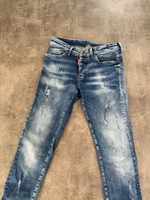 Blå slitna jeans från dsqaured2 - Säljer ett par blå jeans från dsqouard med slitningar och tvättade detaljer för en cool, avslappnad look. Klassisk femficksmodell med knappgylf och röd Levi's-tag vid fickan. Perfekta för dig som gillar streetstyle och vill ha ett par jeans med attityd.