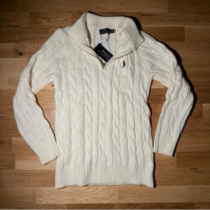 Vit Zip-Knit Polo Ralph Lauren - Kabelstickad vit tröja från Polo Ralph Lauren med hög krage och dragkedja. Tröjan har det klassiska broderade logotypen på bröstet och är gjord i mjukt material. Ribbstickade muddar vid ärmslut och nederkant ger en snygg passform.TVEKA INTE ATT SKICKA EN BUD:) 