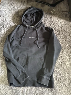 Svart hoodie från Les Deux - Svart hoodie från Les Deux med vit broderad logga på bröstet. Klassisk modell med huva, dragsnören och magficka. Tillverkad i mjukt bomullstyg som är skönt mot huden. Perfekt för en avslappnad och stilren look.
