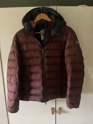 Vinröd pufferjacka från Colmar - Snygg vinröd pufferjacka från Colmar med huva och dragkedja framtill. Jackan har två fickor med dragkedja och Colmar-logga på ärmen. Perfekt för kalla dagar med sin vadderade design och sportiga look.