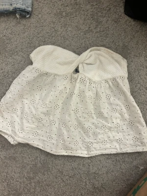 Vit axelbandslös broderad tubtopp - Söt vit axelbandslös tubtopp med twistad byst och broderade blommor över hela tyget. Toppen har en luftig och somrig känsla, perfekt till varma dagar. Materialet känns mjukt och toppen har en bandeau-form.