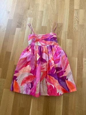 Färgglad blommig sommarklänning - Säljer en kort klänning med tunna axelband och färgstarkt blommigt mönster i rosa, lila, orange och vitt. Perfekt för varma dagar med sitt luftiga tyg och feminina siluett. Klänningen har en dragkedja i sidan och är aldrig använd. Superfin men tyvärr för liten, hittar någon den i S så säg gärna till🤗💞