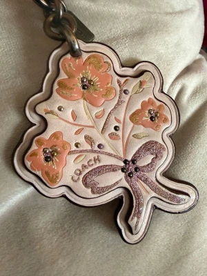 Coach blomformad nyckelring i läder - Superfin blomformad nyckelring från Coach i ljusrosa läder med broderade blommor i persika, guld och glitterdetaljer. Har en glittrig rosett och metallring för enkel fastsättning på väska eller nycklar. Perfekt accessoar för att piffa till din look.