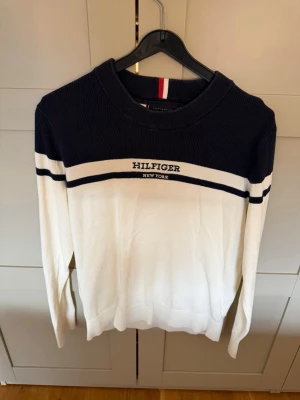 Vit och marinblå Hilfiger tröja - Snygg långärmad tröja från Tommy Hilfiger i vit och marinblå färg. Tröjan har broderad logga på bröstet, rund halsringning och kontrasterande ränder över bröst och ärmar. Perfekt för en stilren och sportig look.