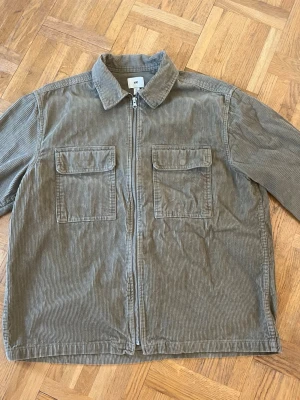 Olivgrön overshirt i manchester - Säljer en olivgrön overshirt i manchestertyg med dragkedja framtill och två stora bröstfickor med lock. Overshirten har en avslappnad passform och klassisk krage, perfekt att slänga över en hoodie eller t-shirt. Inget synligt märke.