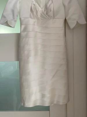 Vit klänning i satin - Elegant vit kortklänning i satin med omlottdesign vid bysten och v-ringning. Klänningen har korta ärmar och ett stilrent, bandat mönster över hela plagget. Perfekt för dig som vill ha en chic och modern look.