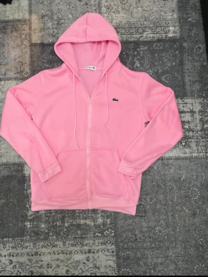 Rosa hoodie från Lacoste med dragkedja - Snygg rosa hoodie från Lacoste med dragkedja framtill, huva med dragsko och klassisk krokodillogga på bröstet. Mjuk och skön bomullsblandning, perfekt för en avslappnad stil. Fickor framtill för extra funktionalitet.