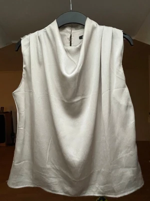 Champagnefärgad satintopp - Snygg ärmlös topp i champagnefärgad satin med draperad halsringning. Toppen har knappar i nacken och ett mjukt, glansigt material!