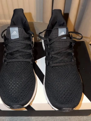 Adidas Ultra Boost svarta sneakers - Svarta Adidas Ultra Boost sneakers med stickad ovandel i mesh och klassiska tre ränder på sidan. Skorna har vita Boost-sulor för extra komfort och svarta snören. Perfekta för en sportig och stilren look. Helt nya