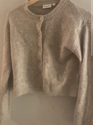 Beige kofta från Monki - Mysig beige kofta från kappahl med rund halsringning och knäppning framtill. Koftan är stickad i ett mjukt material och har långa ärmar samt ribbade muddar vid ärmslut och nederkant. Perfekt för lager-på-lager-stil.