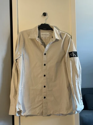 Beige overshirt från Calvin Klein Jeans - Stilren beige overshirt från Calvin Klein Jeans med svarta knappar och klassisk krage. Skjortan har lång ärm och en svart CK-logga på vänster ärm. Perfekt för en clean och modern look. Nypris 1199kr.