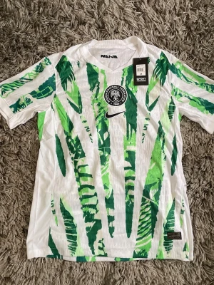Nigeria fotbollströja Nike grön/vit - Säljer en Nigeria fotbollströja från Nike i storlek M. Tröjan är vit med gröna och limegröna mönster, har rund hals och officiellt Nigeria Football Federation-märke på bröstet. Tillverkad i lätt och ventilerande material, perfekt för träning eller match.