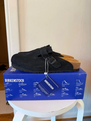 Svarta Birkenstock Boston tofflor - Säljer ett par svarta Birkenstock Boston tofflor i mocka med justerbart spänne och klassisk korksula. Perfekta för dig som gillar stilrena och bekväma skor. Modellen är tidlös och passar till många olika outfits.