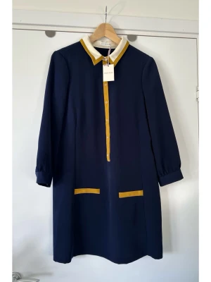 Whistles - helt NY marinblå klänning - storlek 40 - Helt ny stilren klänning från Whistles i en elegant marinblå färg med kontrasterande detaljer i senapsgult och krämvitt. Storlek 40. Klänningen har en klassisk krage och en knappslå framtill med matchande knappar. De långa ärmarna avslutas med manschetter. Klänningen har även dekorativa fickdetaljer framtill i senapsgult. Nypris 1800:-. Helt NY med etikett. 