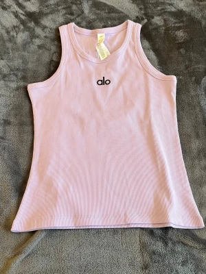 Ljuslila ribbad linnetopp från Alo Yoga - Säljer en ljuslila ribbad linnetopp från Alo med broderad logga framtill. Toppen är ärmlös och har en tight passform med rund halsringning. Perfekt för en sportig och trendig look.