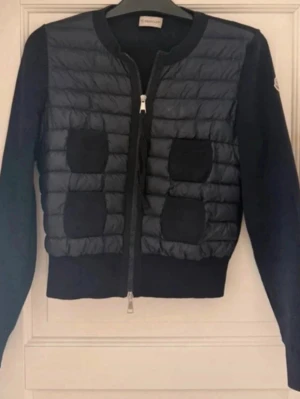 Marinblå cardiganjacka från Moncler - Snygg marinblå cardiganjacka från Moncler med quiltad framsida och stickade ärmar. Jackan har rund halsringning, dragkedja framtill och dekorativa fickor på bröstet. Materialmix av dun och stickat ger en modern look. Storlek S men sitter snarare som XS. Postar dagen eller dagen efter du köper!