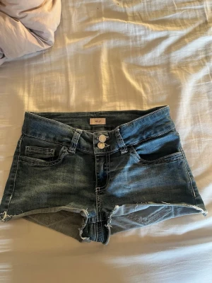 Blå denimshorts från Nelly - Snygga blå denimshorts från Nelly med råa kanter och dubbla knappar framtill. Klassisk femficksmodell med coola broderade detaljer på bakfickorna. Perfekta för varma dagar och enkel att matcha med allt.