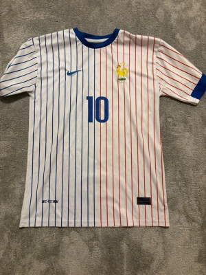 Frankrike Mbappé fotbollströja Nike - Snygg vit fotbollströja från Nike med vertikala blå och röda ränder, blå detaljer vid hals och ärmslut. Nummer 10 och Mbappé tryckt på ryggen, samt franska landslagets emblem på bröstet. Perfekt för dig som älskar fotboll och vill sticka ut på planen.