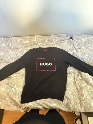 Svart loungewear-set från HUGO - Snyggt svart loungewear-set från HUGO med matchande sweatshirt och byxor. Tröjan har HUGO-logga i vitt med röd ram på bröstet och byxorna har samma logga på benet. Mjukt material och avslappnad passform, perfekt för chill dagar hemma.