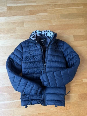 Mörkblå pufferjacka från Superdry - Snygg mörkblå pufferjacka från Superdry med hög krage och vit logotyptext på insidan av kragen. Jackan har dragkedja framtill, justerbar nederkant och quiltad design. Perfekt för kalla dagar och riktigt skön att bära.