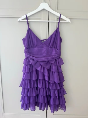 Lila volangklänning med snörning - Superfin lila kortklänning med volanger och smala axelband💜 Klänningen har omlott-detalj framtill och snörning i ryggen för en snygg passform. Tillverkad i ett lätt och luftigt material perfekt på sommaren! Materialet är polyester.