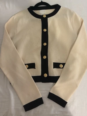 Krämvit kofta med svarta detaljer från H&M - Snygg krämvit kofta med svarta ribbade kanter och guldiga knappar framtill. Koftan har rund halsringning, långa ärmar och dekorativa fickor med svarta detaljer och guldknappar. Perfekt för en chic och stilren look.
