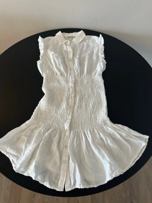 GUESS Vit kort klänning  - Supersöt vit klänning från Guess med knappar hela vägen framtill, krage och volang nertill. Klänningen är ärmlös med små volangdetaljer på axlarna och har smockad midja för en snygg passform. Perfekt för varma dagar!