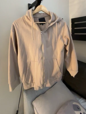 Beige hoodie med dragkedja från lager 157 - Beige hoodie från lager 157 med dragkedja framtill, huva och två fickor. Tillverkad i mjukt material med fleece på insidan för extra komfort. 