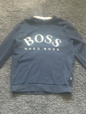Mörkblå sweatshirt från Hugo Boss - Snygg mörkblå sweatshirt från Hugo Boss med stort vitt BOSS-tryck och logga på bröstet. Tröjan har rund halsringning, långa ärmar och ribbade muddar. Storlek XS. 200kr prutat och klart. Bra skick 