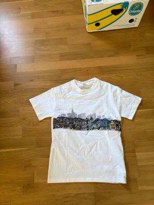 Vit t-shirt med stadsmotiv - Vit t-shirt med tryck av ett färgglatt stadsmotiv över bröstet. Klassisk passform och rund halsringning, perfekt för en avslappnad stil. T-shirten är enkel att matcha med jeans eller shorts.