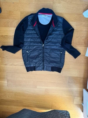 Mörkblåcardigan från Iceland, storlek L - Stilren mörblå Cardigan från Iceland med quiltat mönster och två fickor med dragkedja framtill. Västen har ribbade muddar medans ärmarna är stickade och en diskret röd detalj på insidan av kragen. Samt att Iceland loggan är sydd med gummi plast på ärmen. den är lite stor i storleken Perfekt för lager-på-lager och enkel att matcha.  Storlek L.
