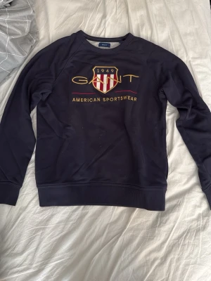 Mörkblå sweatshirt från Gant - Mörkblå sweatshirt från Gant med broderad logga på bröstet. Tröjan har rund hals, ribbade muddar och texten 'American Sportswear' framtill. Perfekt för en stilren look. Använd fåtal gånger, storlek 158/164.