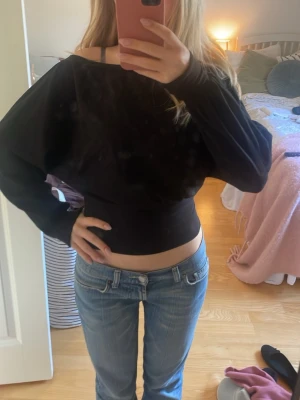 Svart croppad långärmad topp - Snygg svart croppad topp med lång ärm och bred halsringning. Modellen är figurnära över midjan och har en loose passform över axlar och armar. Perfekt för dig som gillar en trendig och avslappnad stil.