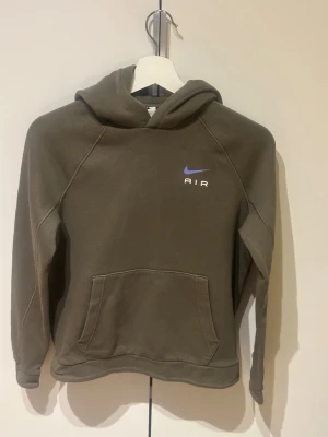 Olivgrön Nike Air hoodie 147-158cm - Säljer en olivgrön hoodie från Nike Air med klassisk känguruficka och huva. Diskret Nike-logga och AIR-tryck i vitt och blått på bröstet. Skön och mjuk bomullsblandning, perfekt för chill dagar eller streetwear-stil.