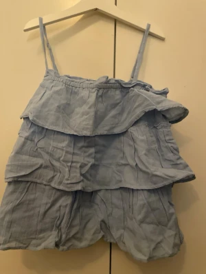 Ljusblå volangig linnetopp - Supersöt ljusblå jumpsuit med tunna axelband och flera lager volanger upptill. Luftigt tyg som känns som bomull, perfekt för varma dagar. Ingen synlig branding. Kort modell med avslappnad passform.