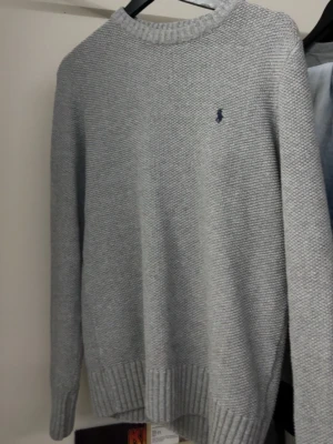 Grå stickad tröja från Ralph Lauren - Snygg grå stickad tröja från Ralph Lauren med diskret blå logga broderad på bröstet. Tröjan har rund halsringning, långa ärmar och ribbade muddar vid ärmslut och nederkant. Perfekt för lager-på-lager och enkel att matcha.