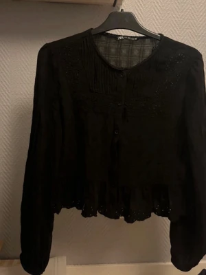 Svart blus med spets från Zara - Svart blus från Zara med knappar framtill, spetsdetaljer och volangkant nedtill. Blusen har lång ärm och rund halsringning. Perfekt för en trendig och stilren look.