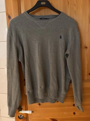 Grå tröja från Polo Ralph Lauren - Klassisk grå tröja från Polo Ralph Lauren med rund halsringning och diskret broderad logga på bröstet. Tröjan har långa ärmar och ribbade muddar vid ärmslut och nederkant. Perfekt för en stilren och enkel look.