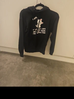 Svart hoodie med katt och text - Svart hoodie med huva och en söt tecknad katt på bröstet. Trycket har texten 'Nope. It looks like bullshit from this angle too' i vitt. Hoodien har klassisk passform och känguruficka framtill. Perfekt för dig som gillar humor och katter.